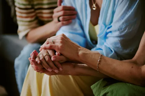Close‑up of one person holding another person’s hands, with a third person resting a hand on their shoulder nearby.