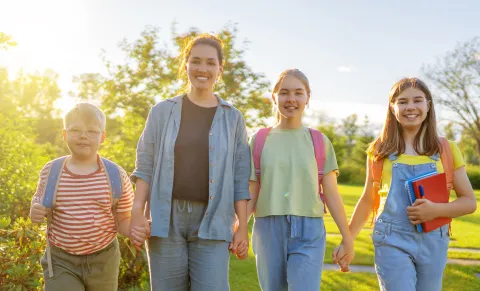 An adult walking with three children with backpacks, holding hands outdoors, with one child carrying a book.