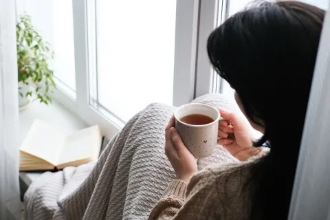 A person wrapped in a blanket sitting by a window, holding a cup of tea, with an open book nearby on the windowsill.
