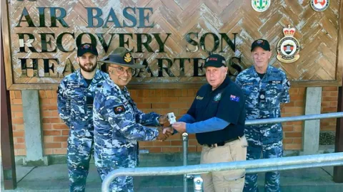A man shaking hands with an Australian Defence Force member in front of a sign that says 'Air Base Recovery Squadron.'