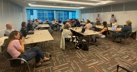 A photo of multiple people sitting in a room with whiteboards.