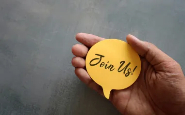 join us-message on yellow note, held in palm of hand - grey background