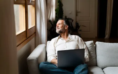 Man sitting on couch, looking out window with a laptop resting on his lap, 