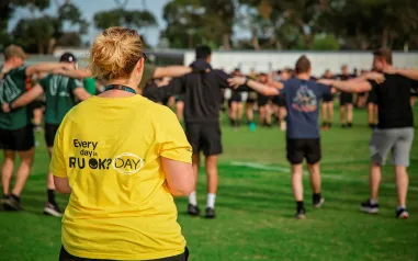 A woman wearing a yellow t-shirt which says Every day is R U OK? Day. A group of people are in the background linking arms.