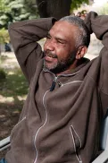 Male sitting on a chair outdoors in a park setting, with arms resting behind their head and trees in the background.