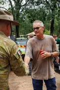 A person in Australian Defence Force  uniform shaking hands with another person outdoors near parked vehicles, with others standing nearby in a bushland setting.