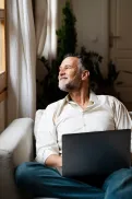 A person sitting on a sofa indoors with a laptop, looking towards a window with natural light.