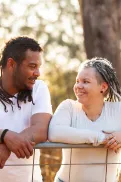 Two people leaning on a fence outdoors and facing each other, with trees and warm sunlight in the background.