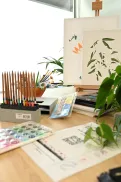 Art supplies arranged on a desk near a window, including coloured pencils, paint and brushes, with a plant and artwork on a stand at the Caboolture Veteran and Family Hub.