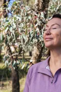 A person standing outdoors among trees with eyes closed, facing the sunlight.