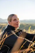 Close-up of a person sitting in tall grass on a hillside, looking upward towards the sky.