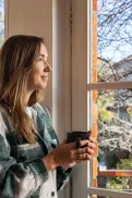 A person standing by a window holding a mug and looking outside.