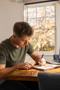A person sitting at a table writing in a notebook, with a laptop nearby.