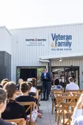 Community gathering outdoors at the Veteran and Family Wellbeing Centre, with a speaker addressing an audience seated in front of the building.