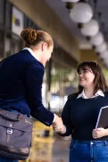 Two people shaking hands in a covered outdoor walkway, with one carrying a shoulder bag and the other holding a folder or notebook.