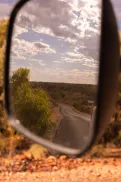 A winding road seen in a vehicle side mirror, with surrounding bushland and trees visible along the roadside under a partly cloudy sky.