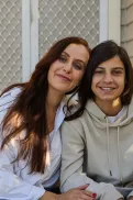 An adult and teenager sitting side by side on front steps, leaning close with an arm around each other.