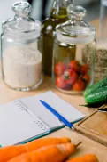 Fresh vegetables, pantry jars and a notebook with a pen arranged on a kitchen bench beside a chopping board and knife.