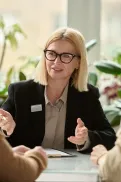 A person in a meeting explaining something to two others seated across a table, with documents in front of them.