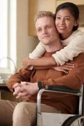 A person in a wheelchair sitting at a kitchen table while another person stands behind with arms around their shoulders.