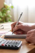 Close-up of a person writing in a notebook with a pen at a desk, with a calculator and laptop nearby.