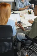 A person in a wheelchair sits at a desk in an office, engaged in conversation with other people around them.