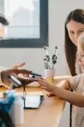 A doctor showing a medical device to a child at a table, with another adult sitting beside them.
