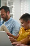 Two adults and a child sitting together on a couch using laptops and a tablet in a home setting.