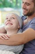 A person sitting on a couch, being hugged from behind by another person resting their arms around their shoulders.