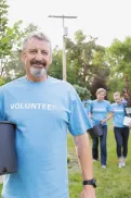 A person carrying a potted tree in a grassy outdoor area, with several volunteers in matching shirts walking behind with plants and tools.