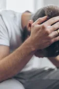 A person sitting on a couch indoors, leaning forward with head in hands.