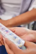 A person holding a labelled weekly medication organiser being passed to another person.