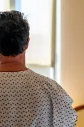 A person wearing a hospital gown sitting on a bed and looking out a window in a hospital room.