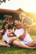 A family sitting on grass outdoors playing together, with one adult lifting a child while others sit nearby in front of a house.