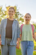 An adult walking with three children with backpacks, holding hands outdoors, with one child carrying a book.