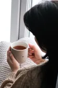 A person wrapped in a blanket sitting by a window, holding a cup of tea, with an open book nearby on the windowsill.