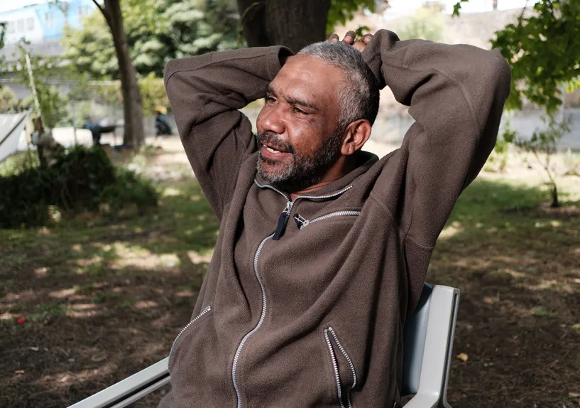 Male sitting on a chair outdoors in a park setting, with arms resting behind their head and trees in the background.