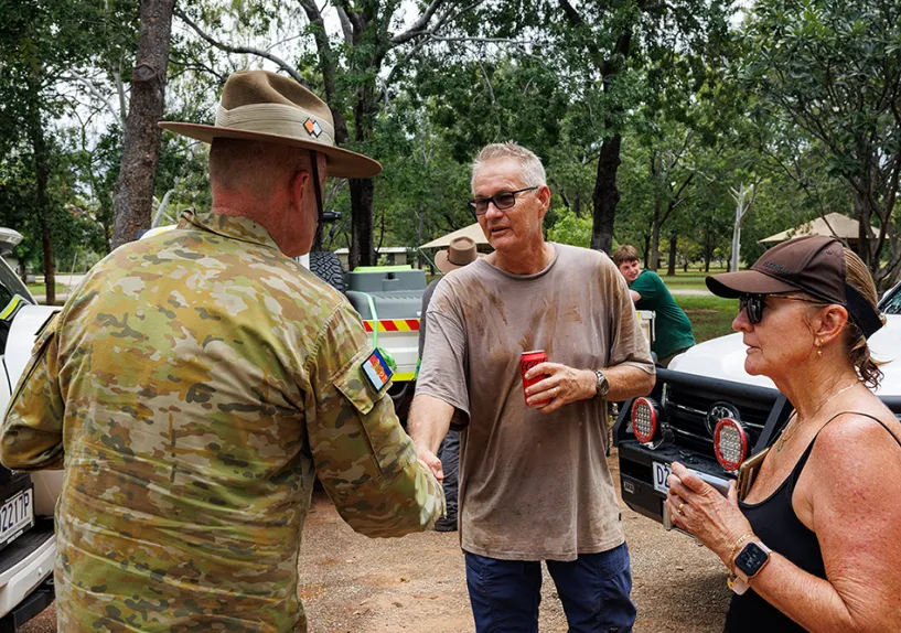 A person in Australian Defence Force  uniform shaking hands with another person outdoors near parked vehicles, with others standing nearby in a bushland setting.