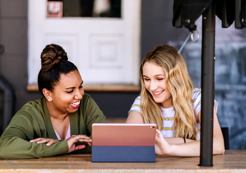 Two adult females sitting together at a table using a tablet in a café or outdoor setting.