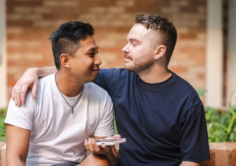Two people sitting close together outdoors, with one person resting an arm on the other’s shoulder while facing each other.