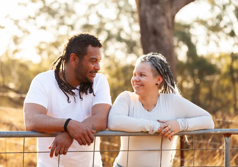 Two people leaning on a fence outdoors and facing each other, with trees and warm sunlight in the background.