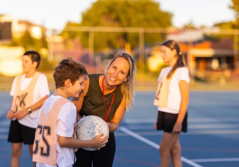 A child holding a ball on a netball court, near umpire and with other children nearby.