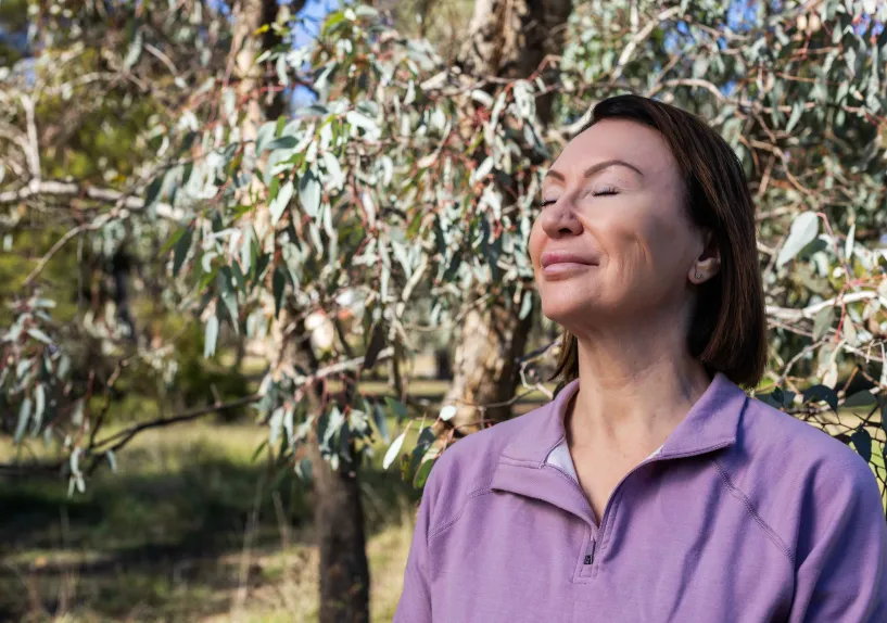 A person standing outdoors among trees with eyes closed, facing the sunlight.