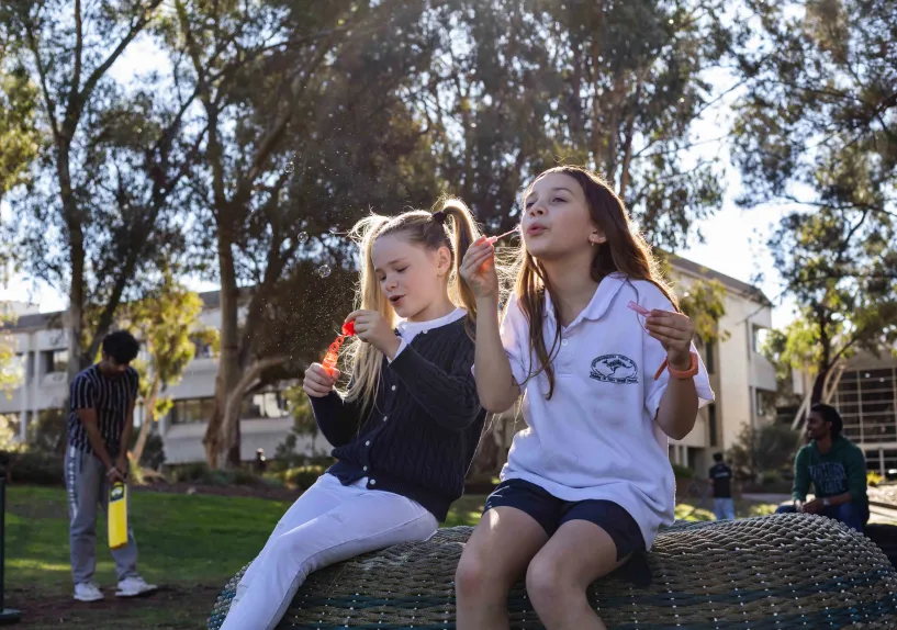 Two young children sitting outdoors in a park, blowing bubbles.