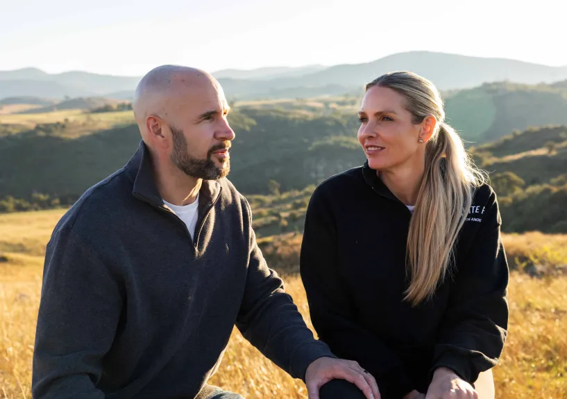Two people sitting together on a hillside, talking, with one resting a hand on the other’s knee.