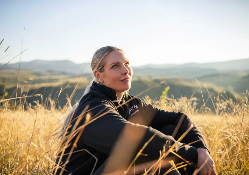 Close-up of a person sitting in tall grass on a hillside, looking upward towards the sky.