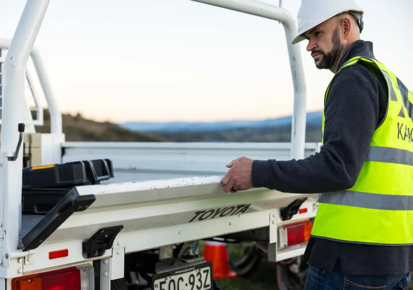 A person wearing a hard hat and high-visibility vest standing at the back of a work vehicle, holding the tray.