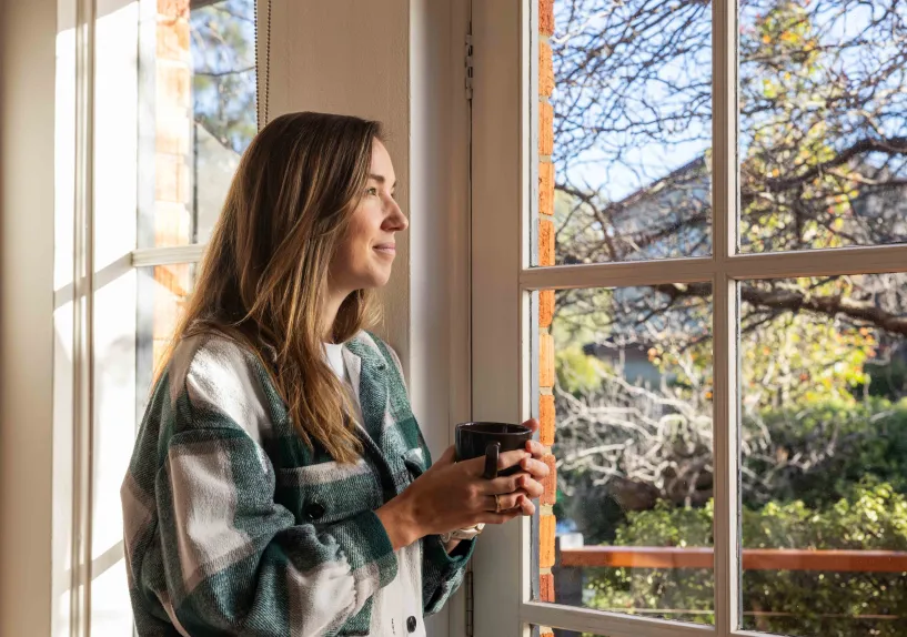 A person standing by a window holding a mug and looking outside.