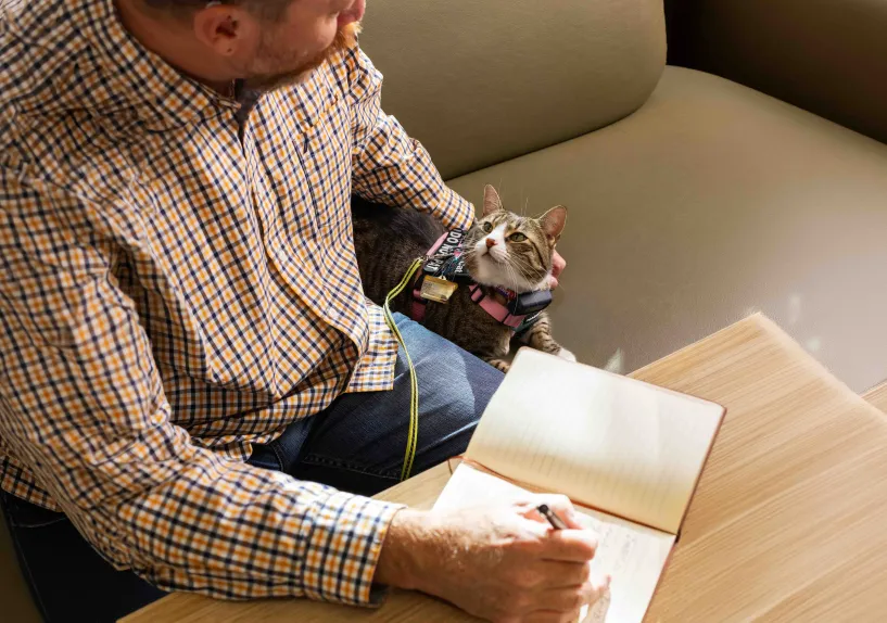 A person writing in a notebook at a table, with an assistance cat beside them.