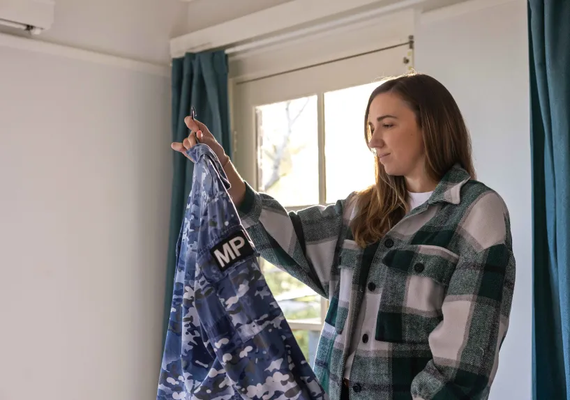 A person holding and looking at their Australian Defence Force uniform on a hanger by a window.